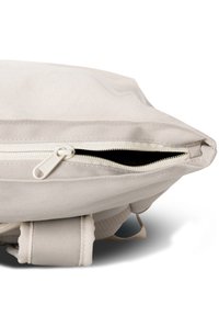 Gros plan d’un sac à dos en tissu beige clair avec une fermeture éclair partiellement ouverte laissant entrevoir le compartiment intérieur et la poignée en tissu en dessous.