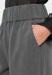 Sivé, textúrované nohavice s širším, elastickým pásom a bočnými vreckami. Tkanina má hladký, jemný pocit s jemným leskom.