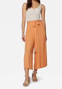 Perzikkleurige plooibroek met wijde pijpen en een strikceintuur in de taille, gecombineerd met een witte kanten tanktop. Zacht materiaal met een lichte, ademende textuur.
