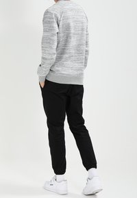 Felpa grigia mélange con maniche raglan abbinata a pantaloni neri a sigaretta. Sneakers bianche con dettagli texture visibili. Sfondo semplice.