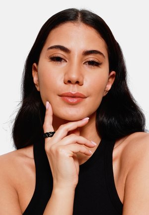 Anello intrecciato nero su un dito, con superficie liscia. L'individuo ha i capelli lunghi e scuri e indossa una maglietta senza maniche nera. Sfondo chiaro.
