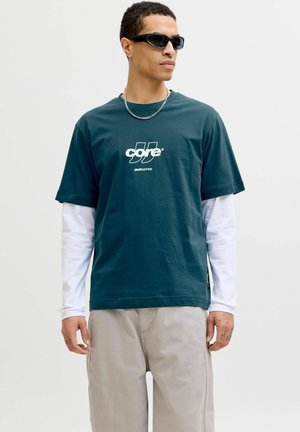 Mørkegrøn bomuld T-shirt med hvidt logo, lagdelt over en hvid langærmet skjorte. Kombineret med lysegrå bukser og solbriller.