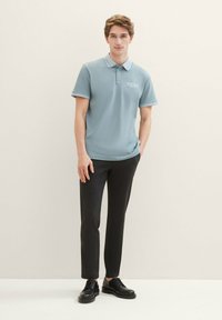 Svetlo modra polo majica z črtastim ovratnikom, kombinirana s temnimi slim-fit hlačami in črnimi veznimi čevlji. Gladka tekstura, sproščen dizajn.