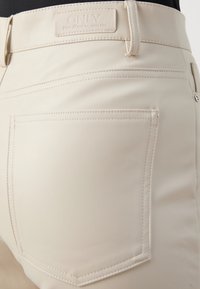 Ljuscremefärgade byxor i konstläder med bakfickor, med ton-i-ton-sömmar och en logotyp vid midjan. Slät textur, figurnära design.