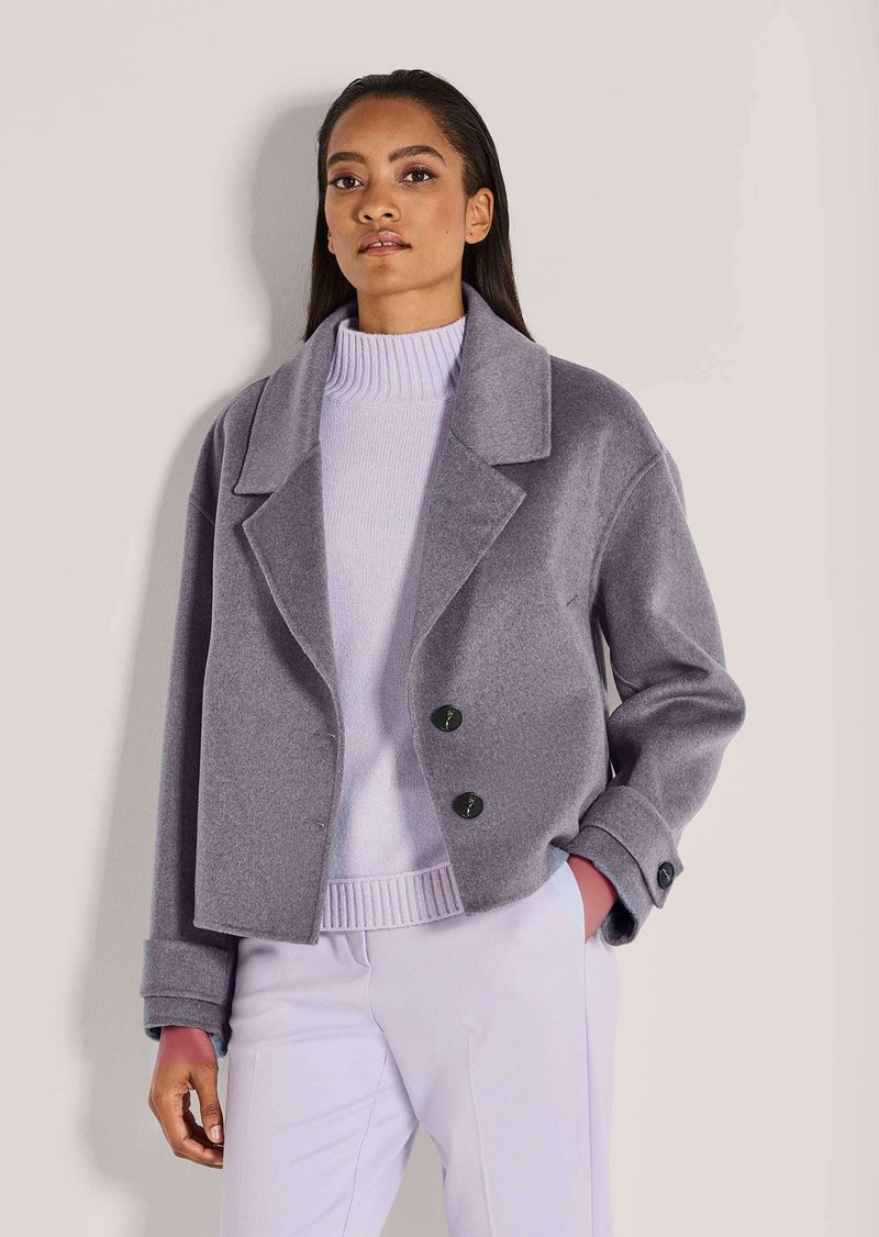 Vrouw draagt een grijze wollen korte jas over een lichtpaarse coltrui en witte broek, staat met één hand in de zak.
