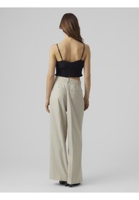 Žena stojící zády, oblečená v černém krátkém topu a béžových kalhotách s vysokým pasem a širokými nohavicemi, s černými podpatky.