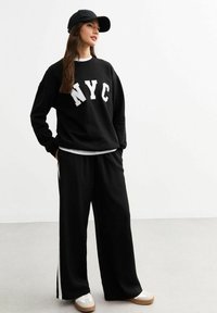 Sudadera negra con letras blancas "NYC", combinada con pantalones de pierna ancha negros con rayas laterales blancas. Llevado con una gorra negra y zapatillas.