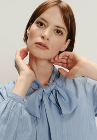 Jasnoniebieska bluzka w paski z wiązanym dekoltem i bufkowymi rękawami. Gładka faktura o lekki połysku i mankiety zapinane na guziki.