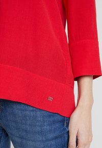 Maglietta rossa a maniche lunghe con maniche a polsino indossata con jeans blu, che mostra un piccolo emblema argento del marchio vicino all'orlo sul lato destro.