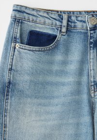 Gros plan sur un jean en denim bleu clair montrant la poche avant avec une doublure de poche bleu foncé, des passants de ceinture et un détail de bouton en métal.