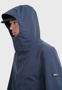 Veste imperméable navy avec un col haut et une capuche. Présente un patch logo sur le côté et une texture lisse et mate. Le design offre une couverture intégrale.