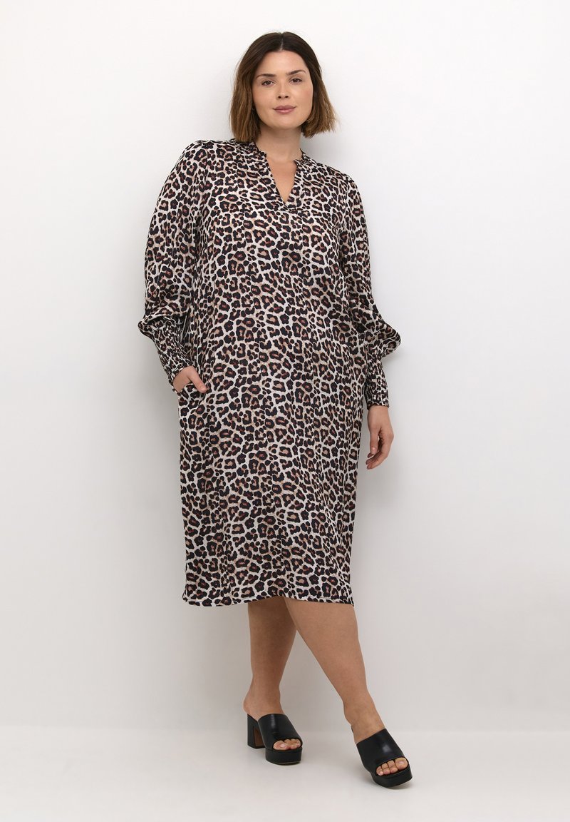 Robe en imprimé léopard avec des manches longues, décolleté en V, tissu fluide, deux poches latérales et longueur au mollet. Portée avec des sandales à talons carrés noires.