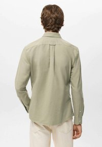 Camisa de manga larga en tela verde claro con dobladillo recto, detalles de botones en los puños y un solo pliegue trasero para facilitar el movimiento.