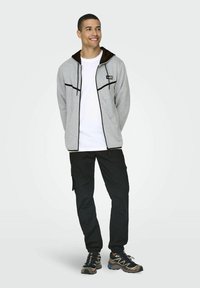 Chaqueta gris con cremallera y detalles en negro, con capucha, un parche de logo, camiseta blanca debajo, pantalones cargo negros y zapatillas de correr multicolores.