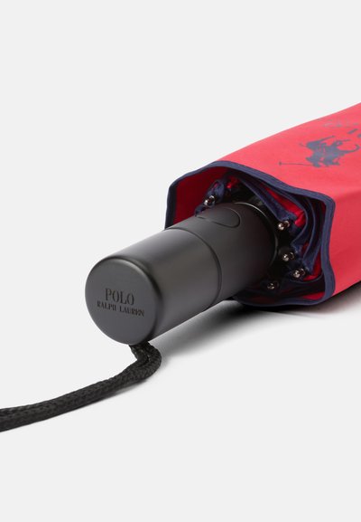 Guarda-chuva dobrável vermelho e azul-marinho com um cabo preto. Possui um logotipo no cabo e uma pulseira trançada. Por dentro, apresenta varetas de metal.