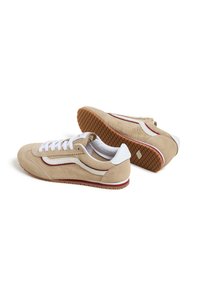 Zapatillas de ante beige con cordones blancos y franjas laterales; suela de goma texturizada con un patrón de banda marrón; detalles rojos en la entresuela.