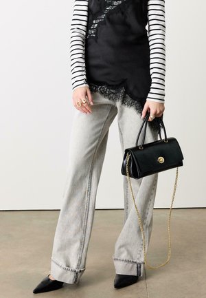 Person trägt ein gestreiftes Langarmshirt unter einem schwarzen Spitzentop, helle graue Jeans mit weitem Bein, schwarze spitze Schuhe und hält eine schwarze Handtasche mit goldener Kette.