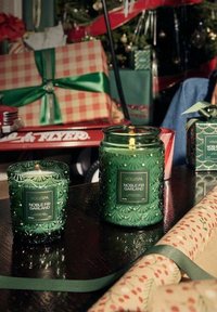 Två gröna glasljus med en texturerad, blommig design, etiketterade "VOLUSPA NOBLE FIR GARLAND," placerade på en mörk träyta.