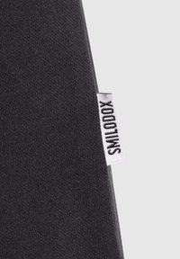 Gros plan sur un tissu noir montrant une petite étiquette blanche avec le nom de la marque "SMOOTOX" en texte noir.
