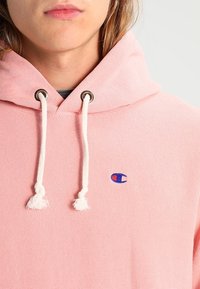 Lichtroze hoodie van gestructureerde stof, met een voorzak, geribbelde manchetten en trekkoorden met witte kwastjes. Het merklogo is zichtbaar.