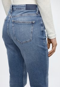 Világoskék farmernadrág magas derékkal, két hátsó zsebbel és márkás bőr címkével. Sima textúra és testre szabott szabás.