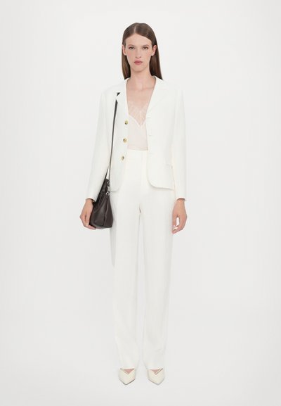 White tailored suit with a fitted jacket, lined pants, and a sheer top. Model holds a small dark bag. Stiletto heels in a nude color.