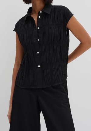 Vrouw draagt een mouwloos zwart gestructureerd overhemd met knopen en een zwarte broek, staand met één hand op haar heup.