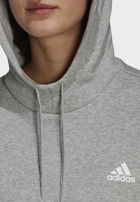 Gros plan d'une personne portant un sweat à capuche Adidas gris clair avec la capuche relevée, doté de cordons de serrage et du logo Adidas sur la poitrine.