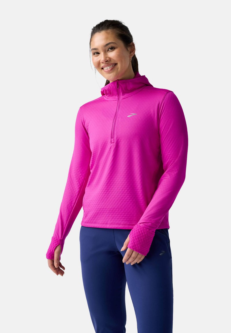 Maglietta aderente rosa a maniche lunghe con colletto a zip, tessuto strutturato, aperture per i pollici e un logo discreto sul petto. Abbinata a pantaloni blu navy.