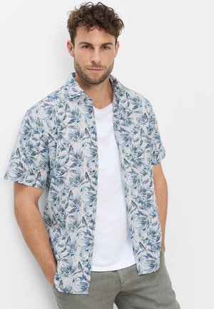 Mann mit lockigem Haar, der ein blau-weißes kurzärmeliges Hemd mit Blattmuster über einem weißen T-Shirt trägt und mit den Händen in den Taschen steht.