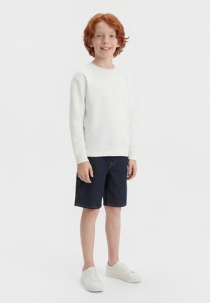 Garçon aux cheveux roux debout et souriant, portant un sweat-shirt blanc, un short sombre jusqu'aux genoux et des baskets blanches, sur un fond clair uni.