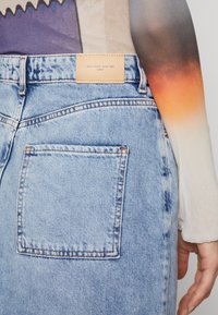 Falda de mezclilla azul clara con un bolsillo trasero cuadrado, que presenta una etiqueta de cuero que dice "BLUE DENIM ANNO 1995 DQILY." Textura suave.