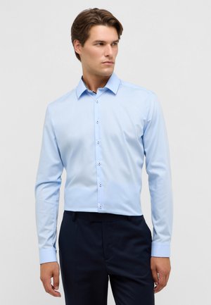 Lichtblauwe overhemd met lange mouwen en een kraag, voorzien van een knoopsluiting aan de voorkant en contrasterende knoopdetails, gecombineerd met donkere broeken.