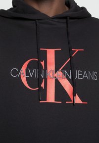 Sudadera negra con una gran logo "CK" rojo y "CALVIN KLEIN JEANS" en texto plateado debajo. Cuerdas con puntas negras. Textura de tela suave.