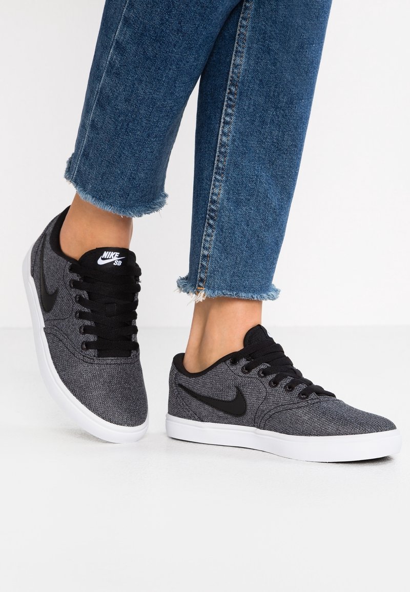 Svarta och grå tygskor med en strukturerad yta, vit gummisula, svarta snören och en liten svart Nike-logotyp på tungan.