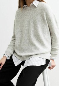 Osoba oblečená v světle šedém svetru přes bílou košili s pruhy a černých kalhotách, sedící na bílé stoličce na prostém pozadí.