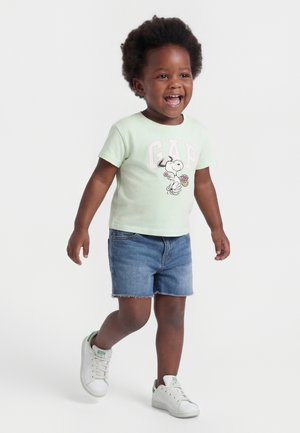Tout-petit aux cheveux courts et bouclés portant un t-shirt Snoopy vert menthe, un short en jean et des baskets blanches, marchant et souriant sur un fond blanc.