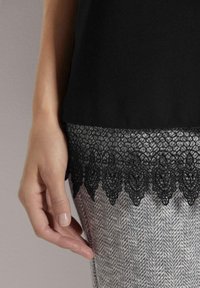 Schwarzes Oberteil mit Spitzenabschluss und filigranen Mustern, über grauem Hahnentritt fabriciert. Glatte Textur und taillierte Passform.