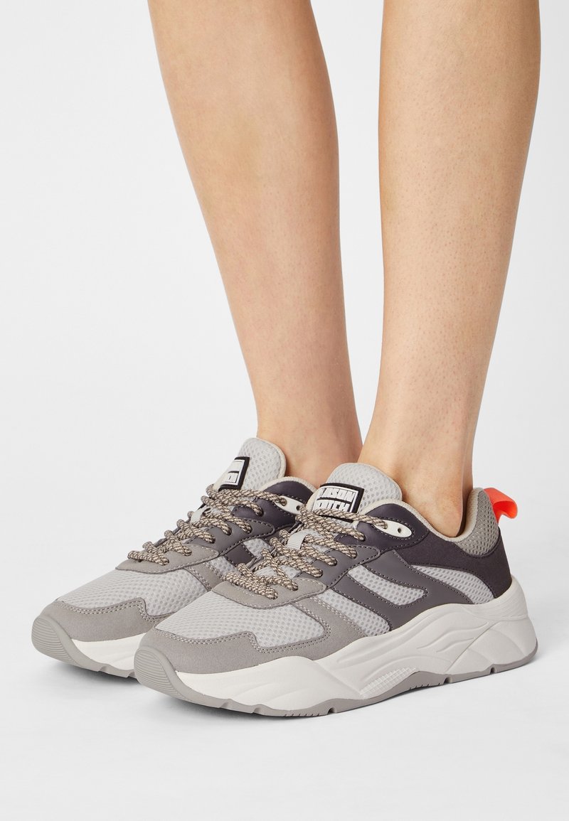 Jambes portant des baskets de sport grises et blanches avec des lacets à motifs et une languette orange au talon, debout sur une surface blanche.