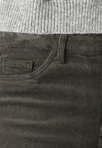 Tmavě olivově zelené kordové kalhoty s výraznými vertikálními pruhy, obsahující malou přední kapsu a standardní poutka na opasek.