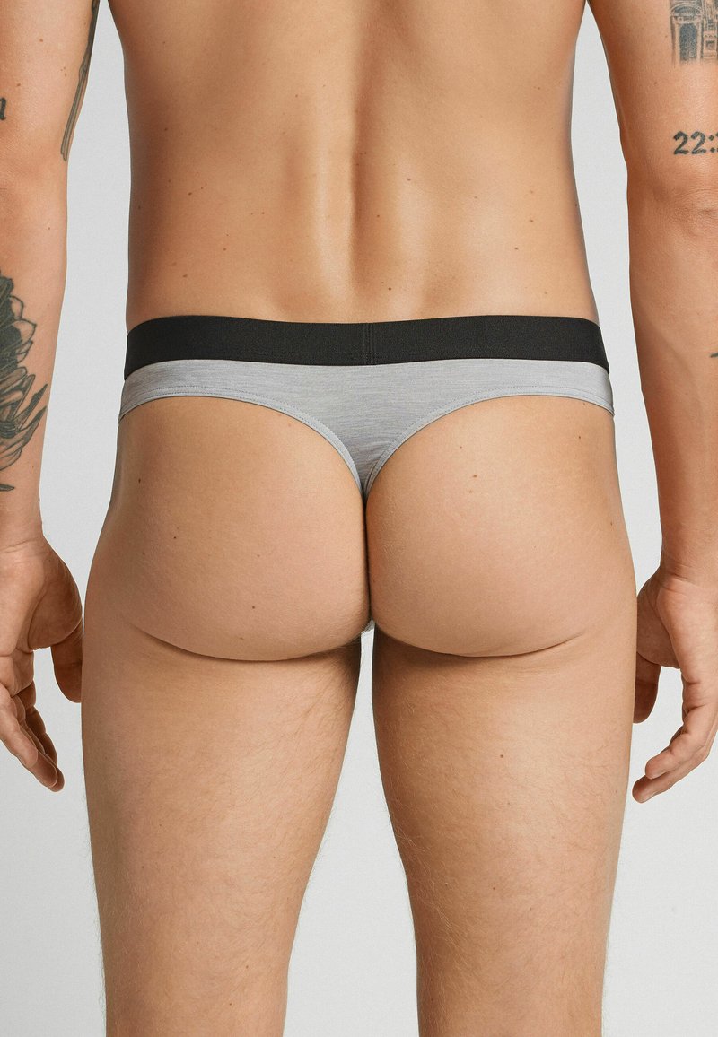 Grijze en zwarte mannen thong onderbroeken met een brede elastische tailleband, een zachte textuur en minimale bedekking aan de achterkant.