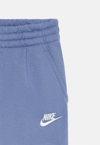 Gaiši zili sporta bikses ar ribotu jostasvietu, sānu kabatām un baltu izbrodētu Nike logo kreisajā pusē. Mīksta auduma tekstūra.