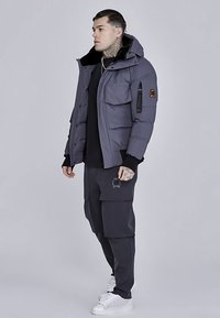 Grijze donzen jas met afneembare capuchon, voorzien van meerdere zakken en een logopatch, gecombineerd met donkere cargobroek en witte sneakers.