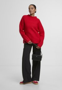 Czerwony, prążkowany sweter z wysokim dekoltem, zestawiony z czarnymi, szerokimi spodniami i butami z czerwonymi akcentami. Trzyma małą czarną torebkę.