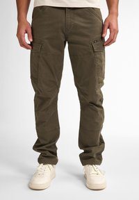 Pantalons cargo vert olive avec plusieurs grandes poches, coupe ajustée et en coton doux. Présente des coutures visibles et une texture usée.