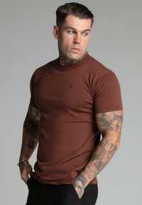 Brown short-sleeve t-shirt with a ribbed round neckline and a small logo. Soft material and fitted design, worn with dark trousers.
