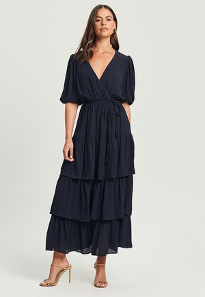 Navy blue tiered maxi dress with short puff sleeves and a V-neck. Features a self-tie waist and flowing, lightweight fabric.