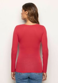 Mujer con cabello largo castaño que se encuentra de frente a una pared blanca, vistiendo una blusa roja de manga larga ajustada y un par de jeans de mezclilla azules.