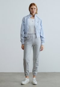 Gaiši zila īsa jaka, balts pieguļošs tops un pelēkas sporta bikses ar elastīgām aprocēm. Modelis valkā baltus sporta apavus ar minimālu dizainu.