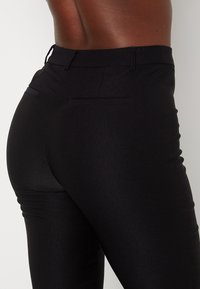 Pantalons noirs fuselés avec une texture lisse, présentant une taille mi-haute, des poches arrière et des lignes épurées, adaptés à diverses occasions.